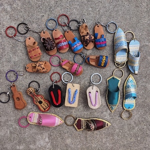 May include: A collection of miniature keychains featuring various designs including leather sandals, flip-flops, guitars, and pointed slippers. The items are crafted from leather, fabric, and metal, showcasing diverse colors like pink, blue, brown, and black.