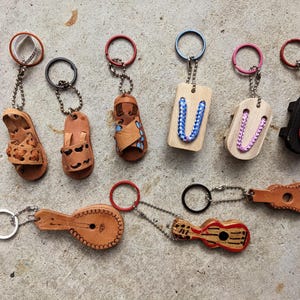May include: A collection of keychains featuring miniature sandals, wooden tags with braided accents, and musical instruments. The keychains are made of leather and wood, with metal key rings and chains. The color palette includes brown, black, red, and blue.