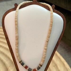 May include: A necklace composed of numerous small, off-white disc-shaped beads strung together. The lower section features a sequence of alternating cylindrical beads in muted red, dark gray, and tan colors, interspersed with the off-white discs.