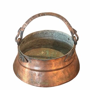 May include: A round copper pot with a wide opening and a curved, hammered metal handle attached by side loops. The exterior has a warm, metallic copper finish, while the interior shows signs of oxidation with green and gray patina.