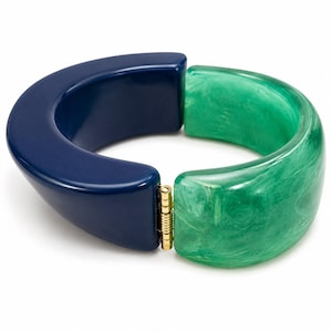 May include: A hinged bangle bracelet featuring two distinct halves. One side is a solid, glossy navy blue resin, while the other side is a translucent, marbled emerald green resin. The two sections are connected by a small, gold-toned metal hinge.