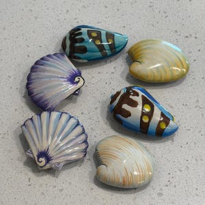 May include: A collection of six decorative seashells arranged in a circular pattern. The shells feature various painted designs, including blue and brown patterns with yellow dots, purple and white ribbed textures, and smooth cream and tan surfaces.