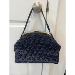 May include: A navy blue, woven handbag with a kiss-lock closure. The bag features a textured, floral-like pattern and a black strap. The frame is a gold color. The bag is hanging on a white door.