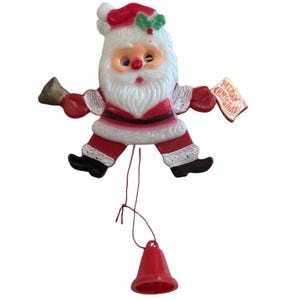 May include: A vintage Santa Claus Christmas ornament. The ornament is red, white, and black, with a red hat and suit. Santa holds a bell in one hand and a sign that says "Merry Christmas" in the other. A red string connects the ornament to a red bell.