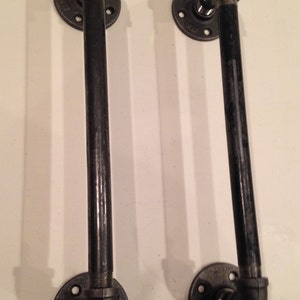 Może przedstawiać: Dwa czarne metalowe uchwyty rurowe z kołnierzami. Uchwyty mają około 30 cm długości i są wygięte pod kątem 90 stopni u góry.