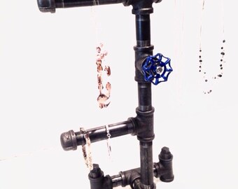 Pode incluir: Suporte de joias de metal preto com design de tubo. O suporte tem uma barra horizontal para pendurar colares e um nível inferior para pulseiras. Uma válvula azul é visível no suporte.