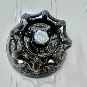 Pode incluir: Uma conexão de tubo de metal preto com uma pega decorativa canelada. A pega tem uma porca hexagonal no centro.