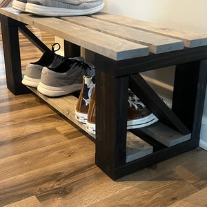 May include: A wooden shoe bench with a gray stained top and black stained legs. The bench has a shelf for storing shoes. There are four pairs of shoes on the bench and shelf, including sneakers and Converse.