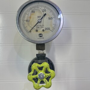 Może przedstawiać: Srebrny manometr z czarną wskazówką i białą tarczą. Manometr wskazuje do 200 psi. Manometr jest przymocowany do czarnego zaworu z zielonym, gwiaździstym uchwytem. Tekst na manometrze brzmi "TUBE-PH. 0.2. CONN-1/4" i "U.S. GAUGE".