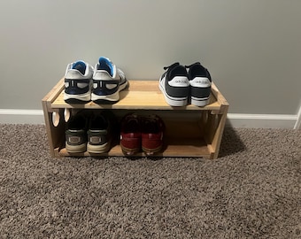 Puede incluir: Un zapatero de madera de dos niveles con varios pares de zapatos. La balda superior contiene zapatillas blancas y azules y zapatillas Adidas negras. La balda inferior tiene pares de zapatos de color oscuro.