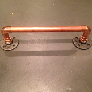 Pode incluir: Um toallero de tubo de cobre com flanges de metal preto. A barra está montada na parede.