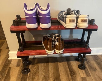 Puede incluir: Un zapatero de dos niveles hecho de madera y tuberías de metal negro. El estante superior tiene dos pares de zapatillas moradas de caña alta y dos pares de zapatos de lona azules con la palabra "DUDE" en ellos. El estante inferior tiene dos pares de zapatillas marrones de caña alta.