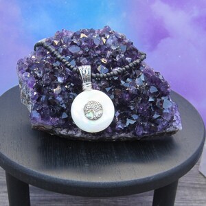 Puede incluir: Un collar de plata con un colgante de ópalo blanco en forma de árbol de la vida en una cadena de cuentas negras. El collar se muestra en un racimo de cristal de amatista púrpura.