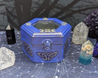 Puede incluir: Una caja de madera azul con tapa de vidrio y un diseño de nudo celta. La caja está rodeada de cristales y flores secas sobre un fondo azul con un patrón de estrellas.
