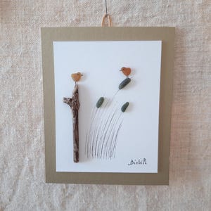 Peut inclure: Une œuvre murale représentant deux petits oiseaux en galets orange sur un fond blanc. Un oiseau est posé sur un morceau de bois flotté vertical, tandis que l'autre est perché sur un roseau dessiné. Des traits d'encre noire forment l'herbe.