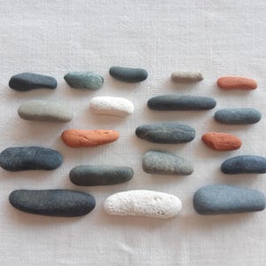 May include: A collection of twenty smooth, elongated river stones arranged in a grid. The stones vary in color, including shades of charcoal gray, light gray, white, and reddish-brown, with some featuring porous textures and others appearing polished.