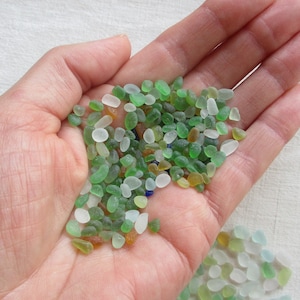 Peut inclure: Une main tenant un tas de petites perles de verre colorées. Les perles sont principalement vertes, blanches et brunes, avec quelques perles bleues mélangées.
