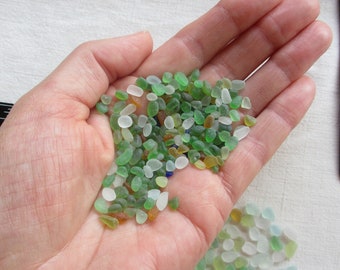May include: A hand holding a pile of small, colorful glass beads. The beads are mostly green, white, and brown, with a few blue beads mixed in.