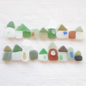 Pode incluir: Uma coleção de peças coloridas de vidro do mar dispostas para se assemelharem a uma cidade costeira em miniatura. As casas, com telhados triangulares e arredondados, são de várias formas e cores, incluindo verde, branco, castanho e azul. O conjunto está sobre um fundo branco.