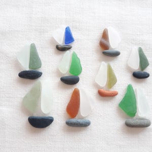 Peut inclure: Huit petites figurines de voiliers disposées sur une surface blanche, chacune fabriquée à partir de morceaux de verre poli par la mer et de petits galets. Les voiles sont en verre blanc, vert, bleu et brun, et les coques sont des pierres.