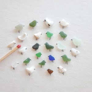 May include: A collection of small, smooth, multi-colored sea glass pieces shaped like birds, each featuring hand-drawn black ink beaks and legs. A single wooden matchstick is positioned on the left for scale, measuring approximately 2 inches long.