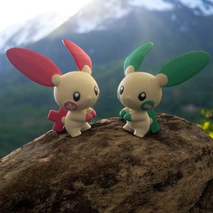 May include: Two small, white, rabbit-like figurines with pink and green ears. The figurines are standing on a large, brown rock with a mountain range in the background.