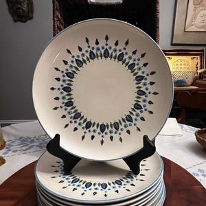 May include: A stack of cream-colored ceramic plates with a blue floral design and a blue rim. The plates are displayed on a black plate stand, showcasing their decorative pattern. The plates are stacked on a wooden surface.