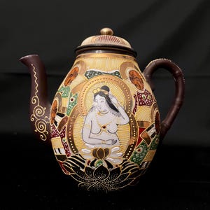 May include: An ornate, hand-painted teapot with a brown handle and spout. The teapot features a central image of a woman with a halo, surrounded by detailed artwork in gold, brown, green, and red. The lid is topped with a gold finial.