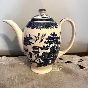 May include: Cream-colored ceramic teapot with a blue Willow pattern design. The teapot features a curved spout, a rounded body, and a matching lid with a small knob. The handle is a curved loop. The blue design includes birds, trees, and buildings.