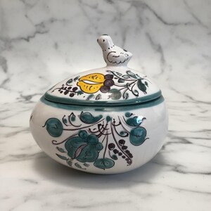 May include: A white ceramic lidded bowl featuring hand-painted floral motifs in teal, yellow, and dark brown. The lid is topped with a small, sculpted white bird figurine. The bowl has a rounded shape with a teal band around the rim.