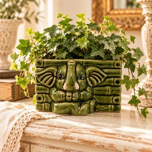 May include: A rectangular green ceramic planter featuring a relief sculpture of an elephant face centered on a bamboo-style background. The planter is filled with a lush green ivy plant with trailing vines extending over the sides.