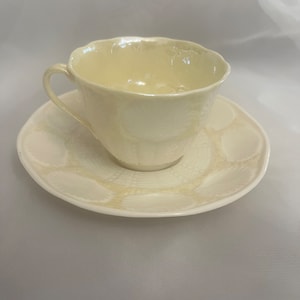 May include: A cream-colored teacup and saucer set. The teacup has a curved handle and a scalloped rim. The saucer is round with a matching scalloped edge. The set is made of a glossy ceramic material.