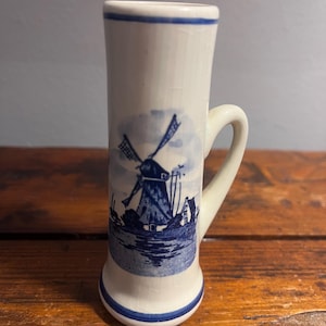 May include: A small, white ceramic shot glass with a blue windmill and landscape design. The handle and rim are also blue. The design is reminiscent of Delftware. The shot glass is on a wooden surface.