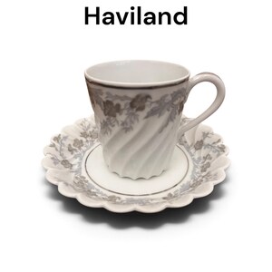 Pode incluir: Um conjunto de ch&aacute;vena e pires em porcelana branca com o texto Valmont by Haviland. A ch&aacute;vena tem um corpo canelado em espiral com um padr&atilde;o floral cinzento e rebordo prateado. O pires tem uma borda recortada com um design floral cinzento.