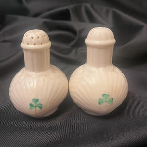 May include: A pair of off-white ceramic salt and pepper shakers featuring a ribbed, rounded body and a painted green shamrock design. One shaker has a perforated top for dispensing, while the other has a solid top with a small central opening.