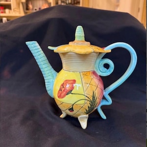 May include: A ceramic teapot featuring a yellow and orange body with a grid pattern, painted red poppy flowers, and green foliage. It has a light blue spout and handle, a yellow lid with a green finial, and stands on three small white feet.