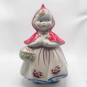 May include: A ceramic cookie jar shaped like a figure wearing a bright red hooded cape. The figure has blonde hair, blue eyes, and holds a small basket. The white dress features red floral accents and a blue trim along the bottom edge.