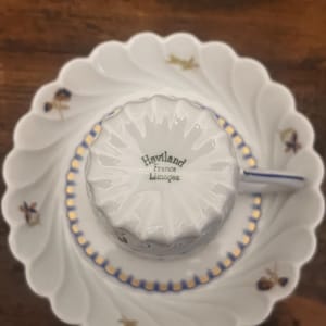 Pode incluir: Um conjunto de ch&aacute;vena e pires em porcelana branca com rebordo recortado. A ch&aacute;vena est&aacute; invertida, exibindo o texto Haviland France Limoges na base. O pires apresenta pequenos motivos florais e um rebordo decorativo em dourado e azul.