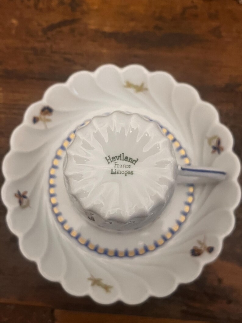 Pode incluir: Um conjunto de ch&aacute;vena e pires em porcelana branca com rebordo recortado. A ch&aacute;vena est&aacute; invertida, exibindo o texto Haviland France Limoges na base. O pires apresenta pequenos motivos florais e um rebordo decorativo em dourado e azul.