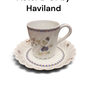 Pode incluir: Um conjunto de ch&aacute;vena e pires em porcelana branca intitulado Aster D'Or by Haviland. A ch&aacute;vena e o pires apresentam uma textura canelada em espiral, um padr&atilde;o de rebordo recortado em azul e dourado e um delicado motivo floral com flores azuis e castanhas.