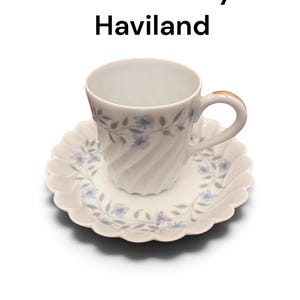 Pode incluir: Um conjunto de ch&aacute;vena e pires em porcelana branca com um delicado padr&atilde;o de videiras florais em azul e roxo. A ch&aacute;vena tem uma textura canelada em espiral e uma asa com rebordo dourado. O texto Gentiane by Haviland est&aacute; impresso acima.