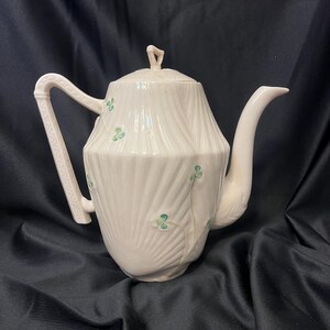 May include: A cream-colored ceramic teapot featuring a vertical ribbed texture and a delicate hand-painted green shamrock pattern. The teapot includes a matching lid with a small finial, a curved spout, and an angular handle with geometric detailing.
