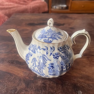 May include: A white and blue ceramic teapot with a lid, featuring a detailed blue willow pattern. The teapot has a curved handle and spout, with gold trim accents. The design includes birds, buildings, and foliage.