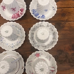 Pode incluir: Seis ch&aacute;venas de ch&aacute; e pires em porcelana branca Haviland Limoges, cada um com um padr&atilde;o floral ou decorativo &uacute;nico. As ch&aacute;venas est&atilde;o invertidas sobre pires recortados, exibindo v&aacute;rias cores, incluindo vermelho, azul, cinzento e dourado.