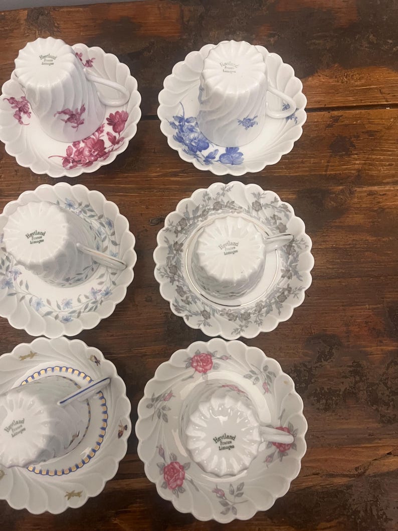 Pode incluir: Seis ch&aacute;venas de ch&aacute; e pires em porcelana branca Haviland Limoges, cada um com um padr&atilde;o floral ou decorativo &uacute;nico. As ch&aacute;venas est&atilde;o invertidas sobre pires recortados, exibindo v&aacute;rias cores, incluindo vermelho, azul, cinzento e dourado.