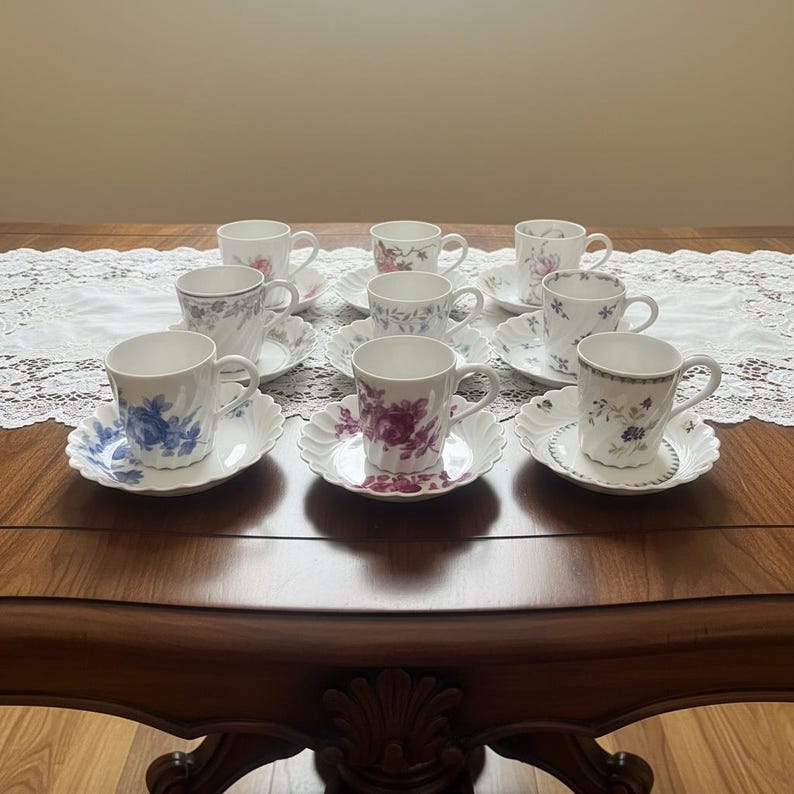 Pode incluir: Um conjunto de seis ch&aacute;venas de ch&aacute; em porcelana branca com pires a condizer, cada uma com padr&otilde;es florais distintos em tons de azul, roxo, rosa e verde. As ch&aacute;venas possuem bordos canelados e pegas, dispostas em duas filas sobre a mesa.