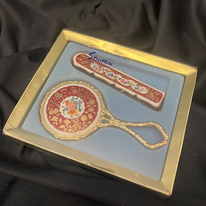May include: A Stratton handbag mirror and comb set in a gold-toned box. The round mirror and rectangular comb feature a red background with a floral pattern and gold accents. The text "HANDBAG MIRROR & COMB SET by Stratton" is printed on the box.