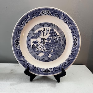 May include: A white ceramic plate with a blue Willow pattern design. The plate features a detailed landscape scene with a bridge, birds, and buildings. The rim has a decorative border. The plate is displayed on a black stand.