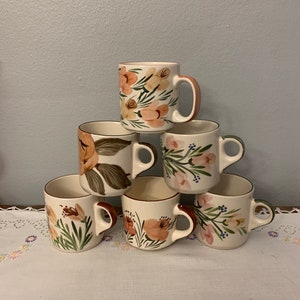 May include: Set of six ceramic mugs with floral designs. The mugs are white with brown, pink, and green floral patterns. The mugs are stacked in a pyramid shape.