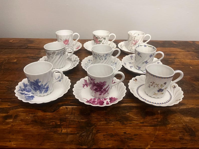 Pode incluir: Um conjunto de oito ch&aacute;venas de ch&aacute; em porcelana branca com pires a condizer, cada uma com um padr&atilde;o floral &uacute;nico em cores como azul, roxo, rosa e verde. As ch&aacute;venas t&ecirc;m uma textura canelada em espiral e bordos recortados.